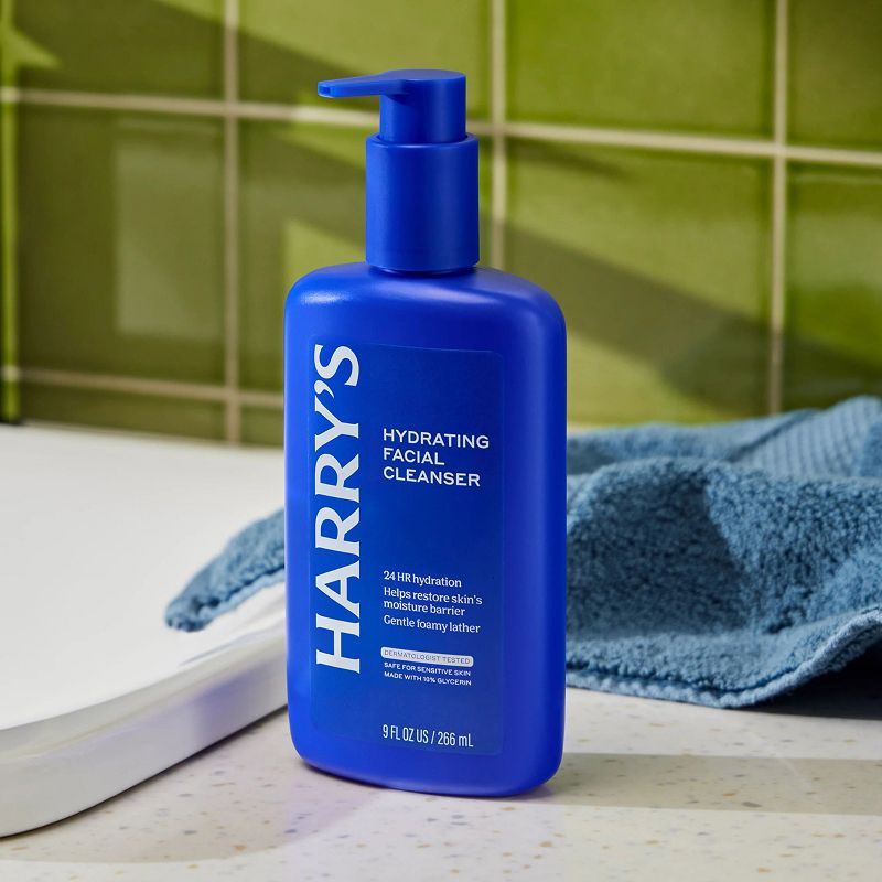 Blue bottle of Harry's hydrating facial cleanser on a bathroom counter with a green tiled wall in the background.